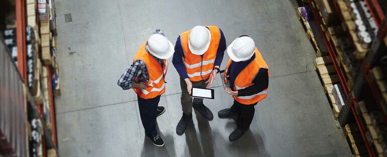 Three people wearing orange safety vests and white hard hats stand in a warehouse, looking at a tablet. The image is taken from above, showing shelves on either side and a concrete floor.