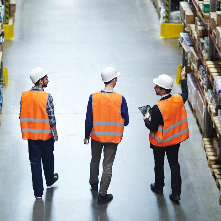 Three workers wearing orange safety vests and white hard hats walk down an aisle in a warehouse, surrounded by shelves stocked with various boxes and products. One person is holding a clipboard.