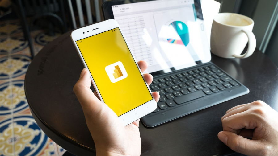 A person holds a smartphone displaying the Power BI logo, while a tablet with a keyboard on a round table shows data charts; there is also a white coffee mug beside the tablet.