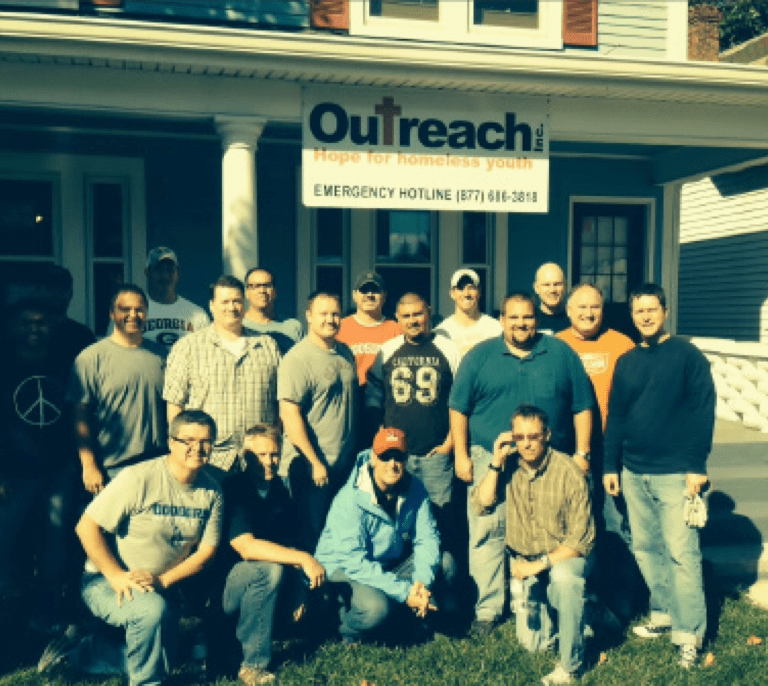 A group of men pose and smile in front of a white house with a sign reading “Outreach: Hope for Homeless Youth. Emergency Hotline (877) 614-3818.” The group stands on grass under bright sunlight.