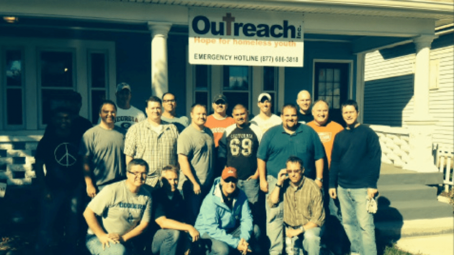 A group of men pose and smile in front of a white house with a sign reading “Outreach: Hope for Homeless Youth. Emergency Hotline (877) 614-3818.” The group stands on grass under bright sunlight.