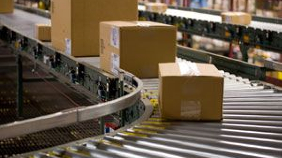 Several cardboard boxes move along a curved conveyor belt in a warehouse or distribution center, with shelves and additional packages visible in the background.