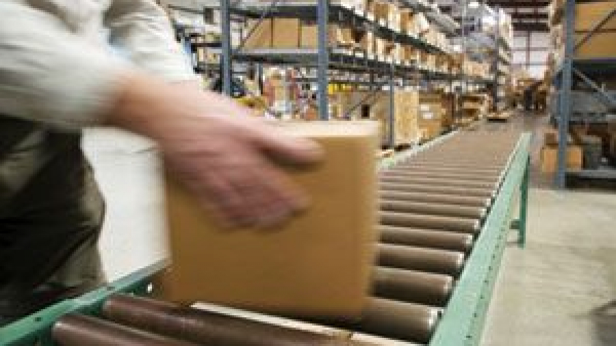 A person places a cardboard box on a conveyor belt in a warehouse filled with shelves of packed boxes.