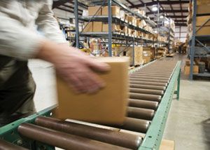 A person places a cardboard box on a conveyor belt in a warehouse filled with shelves of packed boxes.