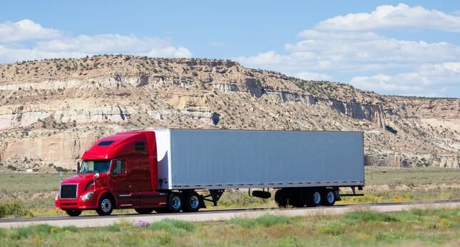 A bright red semi-truck with a long, gray trailer drives along a highway in a desert landscape, with rocky hills and a partly cloudy sky in the background.