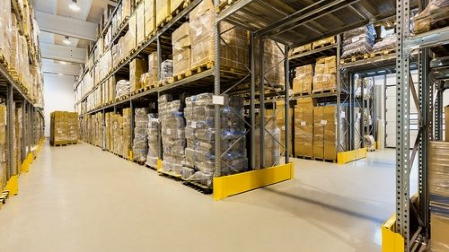 Warehouse interior with tall shelves stacked with cardboard boxes and packages. The floor is clear and clean, with wide aisles for movement. The area is well-lit with overhead lights.