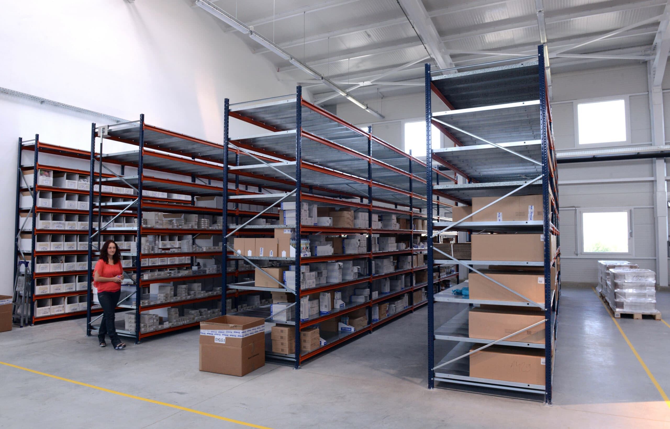 A woman walks through a spacious warehouse with tall metal shelves holding various boxes and items. The area is well-lit with high ceilings and large windows. Some boxes are stacked on the floor.