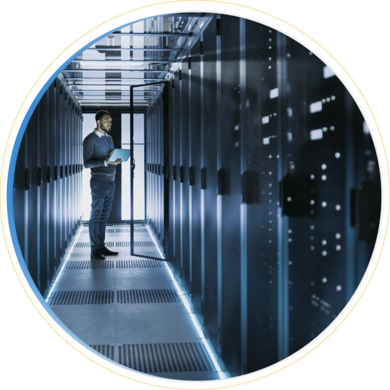 A person stands in a narrow, modern server room holding a laptop, surrounded by tall racks of servers with blue LED lights illuminating the corridor, focused on storage management.