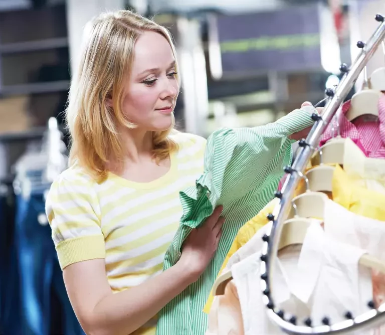 A woman with blonde hair wearing a yellow and white striped shirt is shopping for clothes, examining a green striped shirt on a rack—perhaps inspired by the store’s efficient multi echelon inventory optimization.