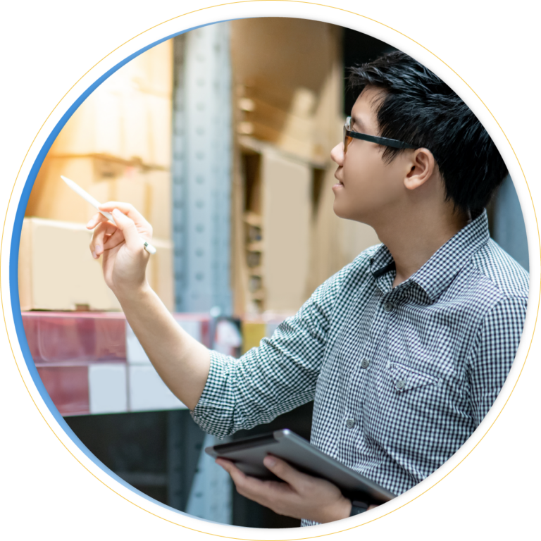 A person wearing glasses and a checkered shirt is holding a tablet and pointing at boxes on shelves, possibly checking inventory or involved in inventory balancing in a warehouse.