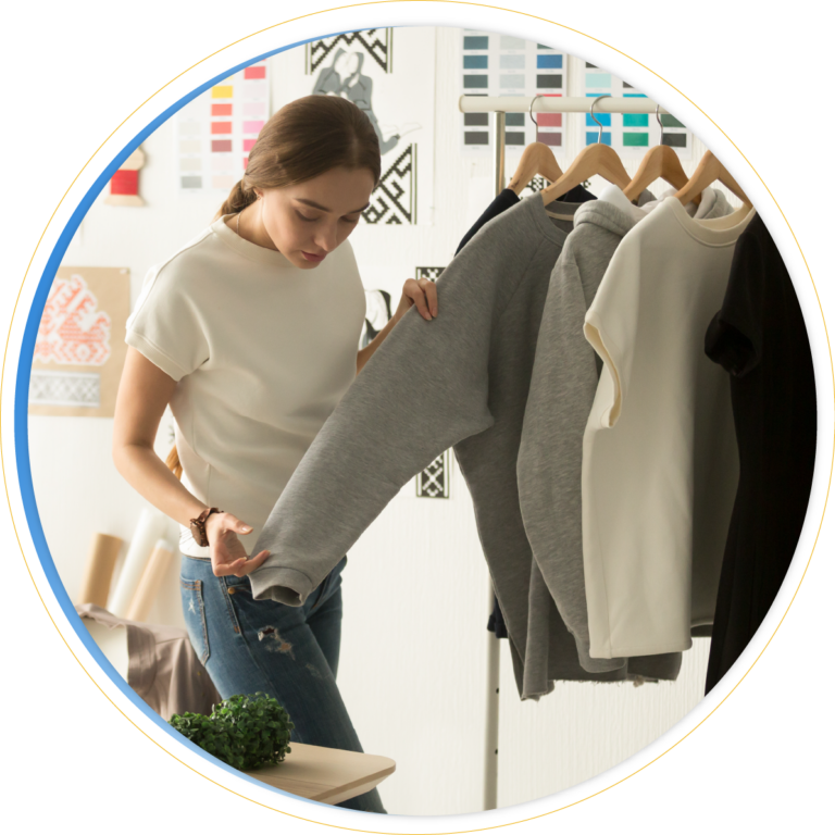 A woman examines a gray sweatshirt from a diverse product assortment on a clothing rack, surrounded by various sweaters in a bright room decorated with color palettes and textile samples.