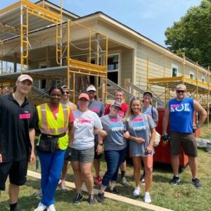 A group of volunteers stands and smiles in front of a house under construction, surrounded by scaffolding, on a sunny day. Most are wearing matching shirts and work gear, highlighting their shared commitment to community impact.