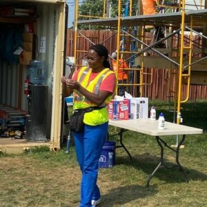 A woman in safety gear stands outside near a table with supplies, checking her phone. Construction workers and scaffolding are visible in the background, highlighting the community impact of their work on a sunny day.