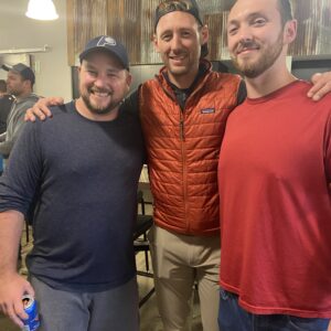 Three men smile and pose together indoors, reflecting community impact. The man on the left wears a blue cap and shirt, holding a drink. The center wears a red vest and cap, while the right sports a red shirt. Others and industrial decor appear in the background.