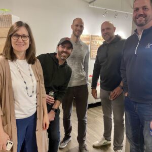 Five adults, four men and one woman, stand together indoors in a casual setting, smiling at the camera. They wear casual clothes in a modern room with a wooden floor, reflecting their shared sense of community impact.