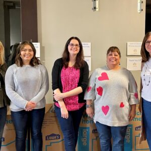 Five women stand smiling in front of stacked Lowe’s Home Improvement boxes, highlighting their community impact. Some boxes display printed signs, and the group appears to be indoors, possibly at an event or workplace.