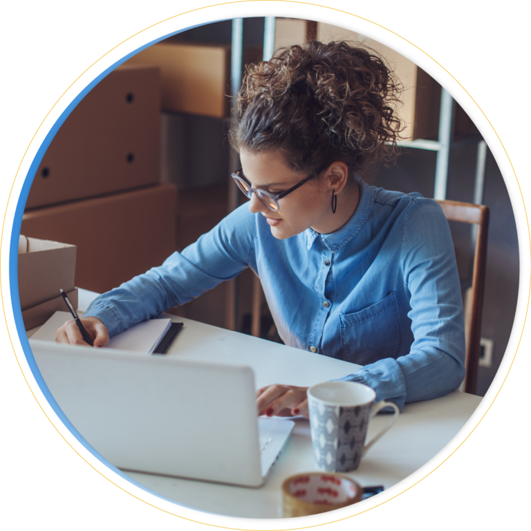 A woman with curly hair and glasses is writing in a notebook at a desk with a laptop, mug, and boxes—perhaps reviewing merchandise planning solutions. She is wearing a blue button-up shirt and appears focused on her work.