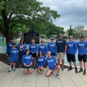 A group of twelve people in matching blue HOPE shirts stand and kneel together on a paved walkway near a canal, showcasing their community impact amid trees, a bridge, and a historical marker sign in the background.