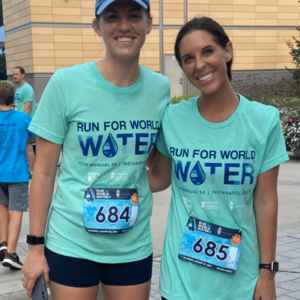 Two women wearing matching “Run for World Water” T-shirts and numbered race bibs smile outdoors at a race event, showcasing community impact as fellow participants and a building appear in the background.