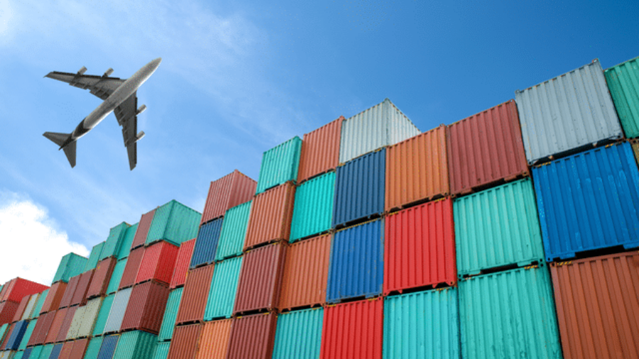 A commercial airplane flies above a large stack of colorful shipping containers under a clear blue sky.