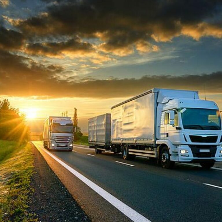 Two large white trucks drive on a highway through green fields at sunset, with dramatic clouds and sunlight shining in the sky.
