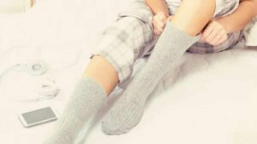 Person sitting on a bed wearing plaid pajamas, pulling up gray socks. A pair of headphones and a smartphone are lying nearby on the bed.