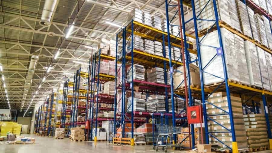 Large warehouse interior with high shelves stacked with various pallets and boxes, organized in rows. The space is well-lit with overhead lighting, and there are carts and packing materials on the concrete floor.