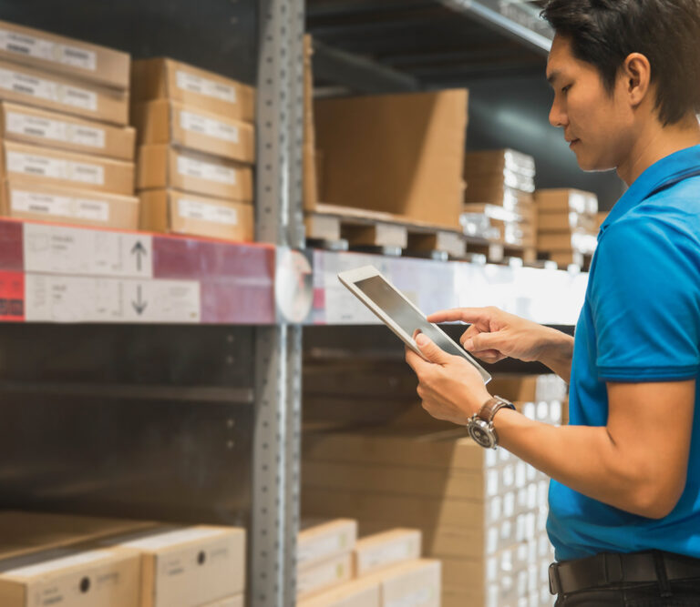 A man in a blue polo shirt uses a tablet while standing in a warehouse, surrounded by shelves filled with organized boxes typically found in micro-fulfilment centers.