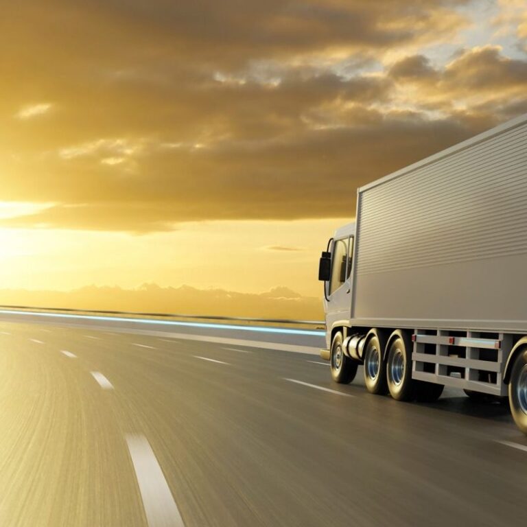 A large white semi-truck drives on a wide, empty highway at sunset, with golden sunlight reflecting off the road and dramatic clouds in the sky.