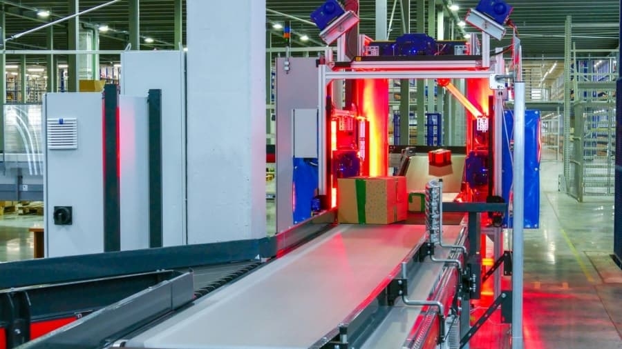 Conveyor belt system in a warehouse with packages being scanned under red lights. The automated machinery and shelving units are visible in the background, indicating a high-tech logistics or sorting facility.