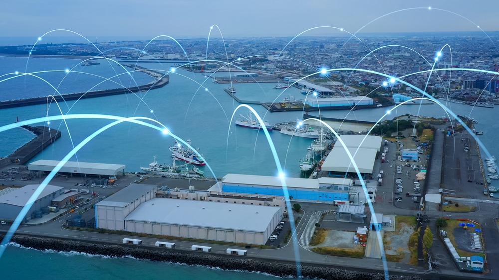 Aerial view of a port city with shipping docks and warehouses, overlaid with digital lines and points representing data communication or a responsive supply chain network.