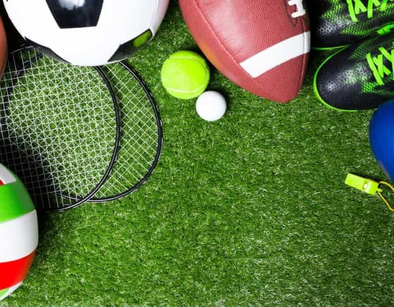 Various sports equipment, including soccer ball, football, volleyball, tennis ball, baseball, badminton rackets, shoes, whistle, and boxing glove, arranged on green artificial grass.