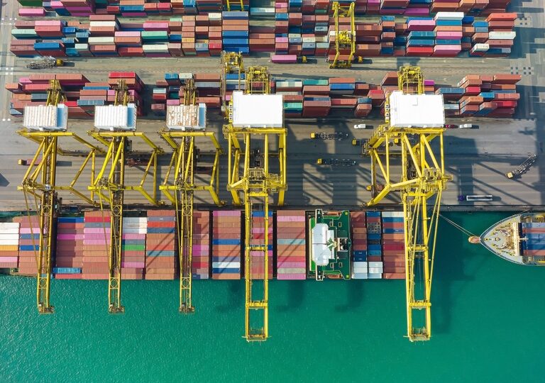 Aerial view of large yellow cranes loading and unloading colorful shipping containers onto cargo ships docked at a busy port, with turquoise water alongside the pier.