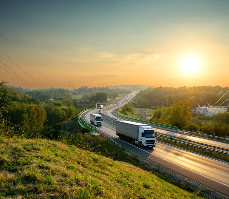 Two semi-trucks drive along a winding highway through green hills at sunrise, the clear sky casting a warm glow—an ideal setting to illustrate a transportation management system business case. Power lines and trees border the serene road.