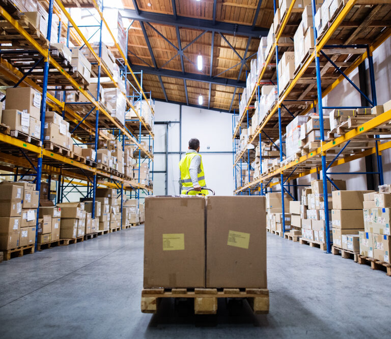 A person in a high-visibility vest stands in a warehouse surrounded by tall shelves stacked with cardboard boxes, with two large ab701 boxes on a pallet jack in the foreground.