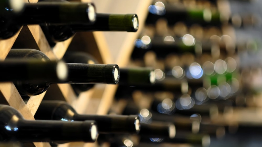 A close-up of wine bottles stored horizontally in a wooden rack, with the bottle necks facing outward. The background shows more wine bottles out of focus.