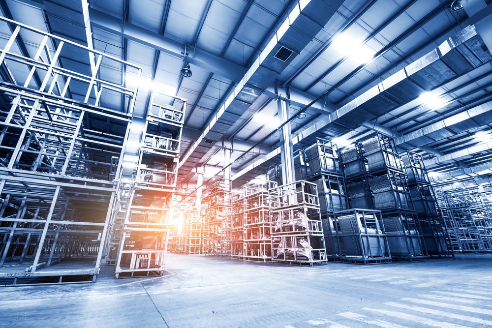 Large, modern warehouse interior with tall metal storage racks and wide aisles, optimized for slam line automation. Bright lighting and a warm sun flare fill the facility, creating a clean, organized atmosphere.