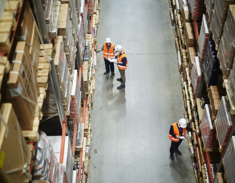 Overhead view of three workers in safety vests and helmets checking shelves and inventory in a warehouse aisle filled with stacked boxes and goods.