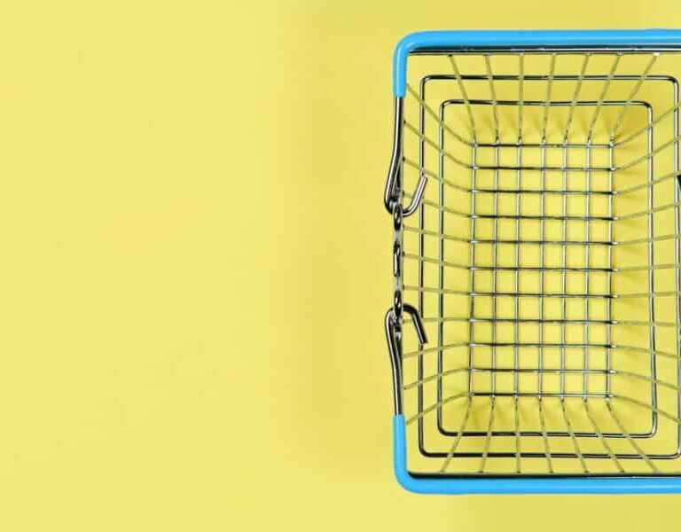 A small, empty metal shopping basket with blue handles is viewed from above against a yellow background, positioned to the right side of the image.