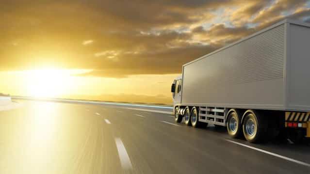 A large white semi-truck drives down a highway at sunset, with golden sunlight illuminating the road and sky, creating a dramatic and serene atmosphere.