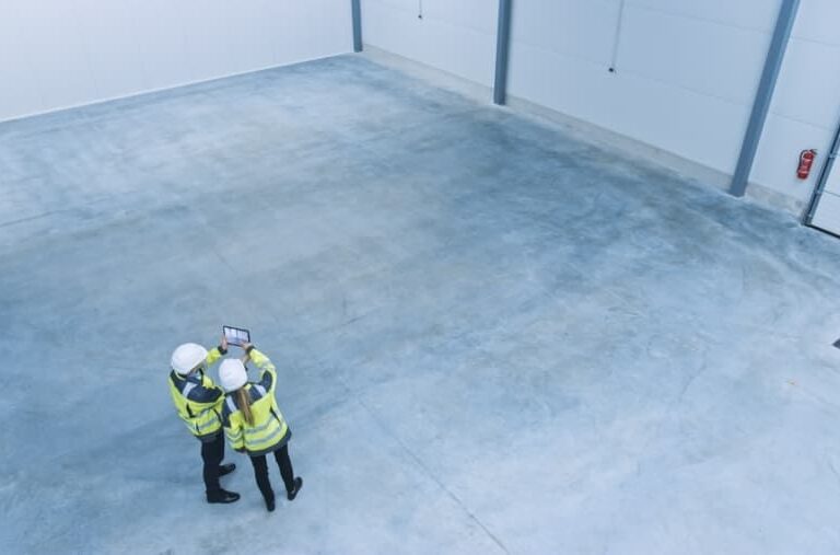 Two people in safety vests and helmets stand in an empty, clean warehouse, looking at a tablet. The space has gray concrete floors and white walls, with a large garage door visible on the right.