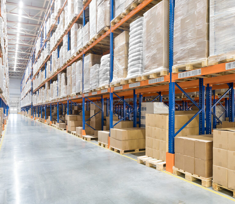 Large warehouse interior with tall blue and orange metal shelving racks stacked with cardboard boxes and wrapped pallets on both sides of a wide, polished concrete aisle. No people are visible.