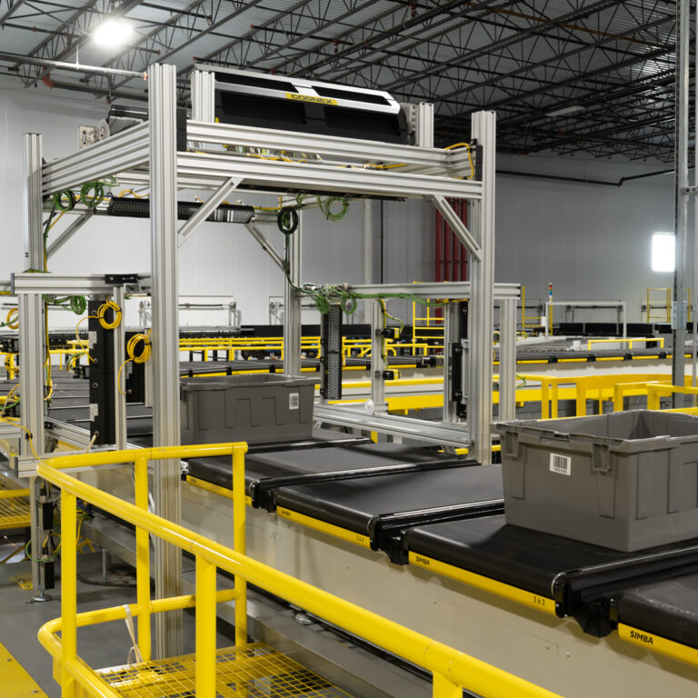 A large industrial warehouse features automated conveyor belts with gray plastic bins and a metal framework, bordered by yellow safety railings. The scene is clean and brightly lit.