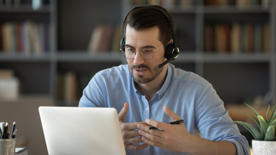 A man wearing glasses and a headset sits at a desk, gesturing as he discusses Microsoft’s Sales Copilot during a video call on his laptop. A bookshelf is visible in the background.