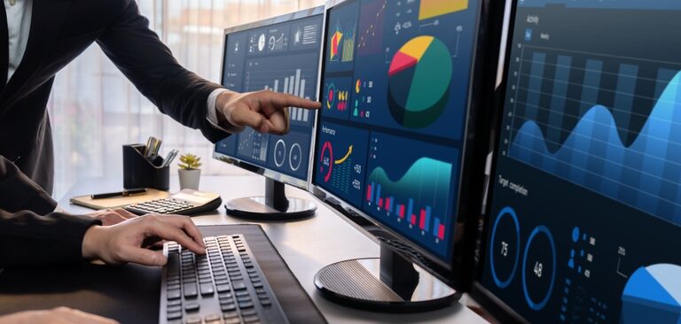 Two people analyze colorful financial graphs and charts powered by Microsoft Business Solutions on three large monitors in a modern office. One types on a keyboard while the other points at the screen, discussing data trends.