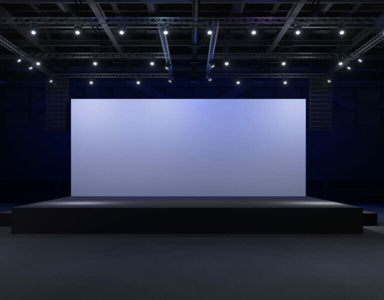 A large, empty stage with a wide, blank screen is lit by overhead spotlights in a dark, spacious auditorium—perfect for unveiling insights from a WMS Implementation Case Study.