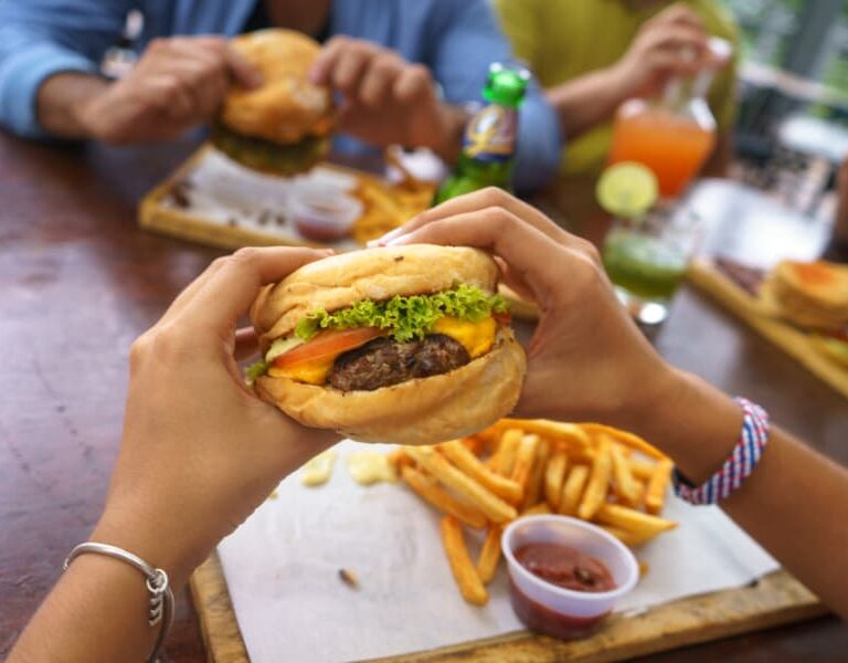 A person holds a burger with lettuce, tomato, and cheese over a tray of fries and ketchup, while others eat burgers and drink beverages at a wooden table.
