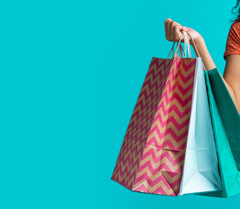 A person wearing an orange polka dot shirt holds several colorful shopping bags against a bright turquoise background. Only the arm and part of the torso are visible.