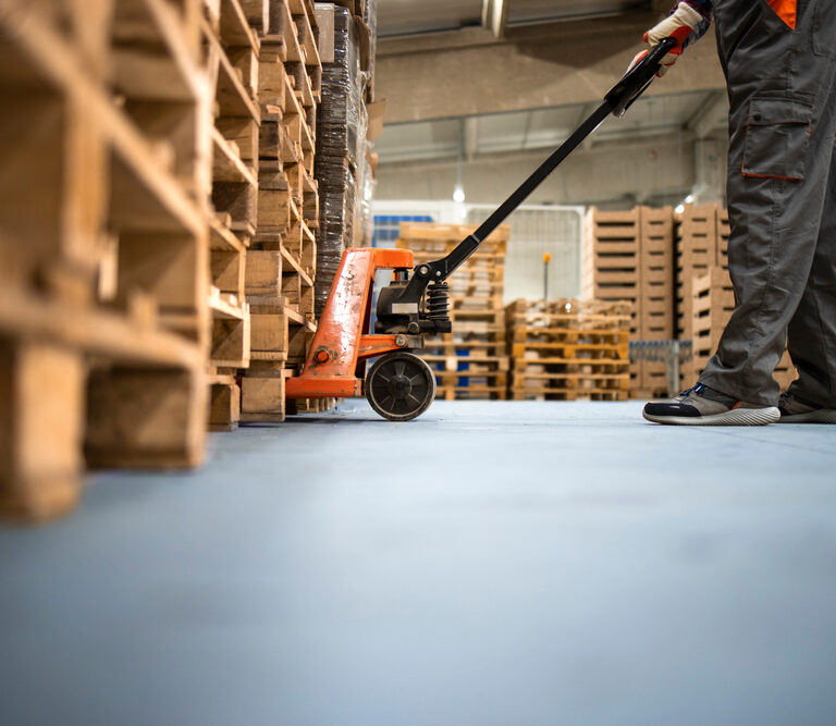 A worker in gray pants and shoes uses a hand pallet jack to move wooden pallets inside a warehouse with stacks of pallets in the background.