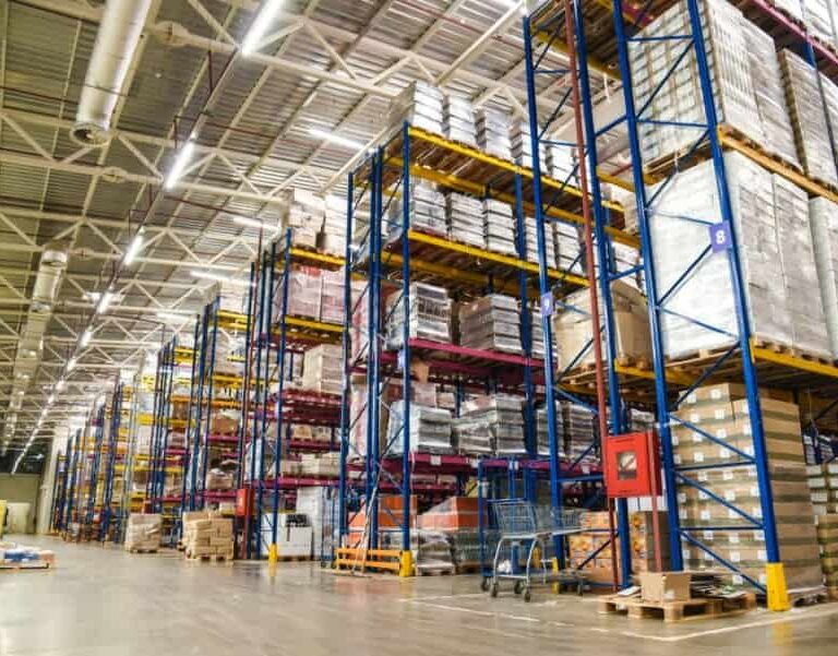 Interior of a large warehouse with tall shelves stacked with various pallets and boxes, organized in rows. The floor is clear, and bright overhead lights illuminate the spacious area.